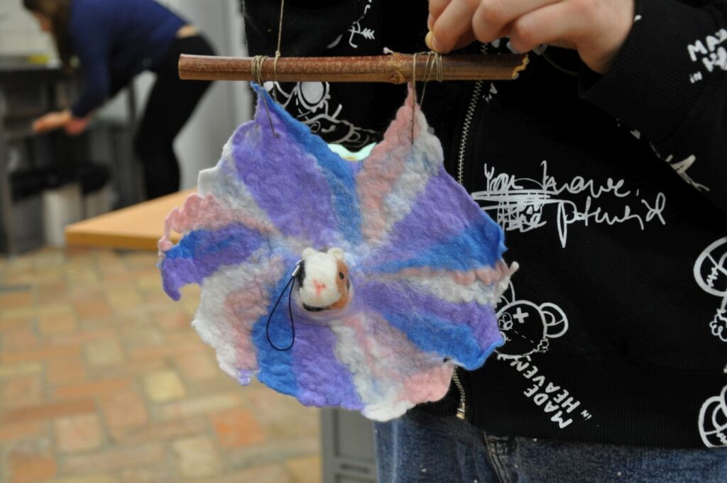 Filcowana ozdoba w kształcie kwiatu w odcieniach fioletu i błękitu  zawieszona na drewnianym patyku, w środku filcowa głowa małej myszy (kliknięcie powiększy zdjęcie).