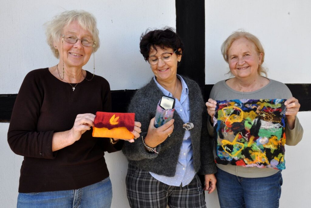 Trzy uczestniczki warsztatów stoją obok siebie i prezentują wykonane przez siebie filcowane prace – etui oraz kolorową tkaninę z wełny (kliknięcie powiększy zdjęcie).