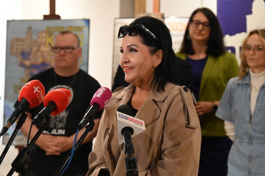 Kobieta w beżowym płaszczu przemawia podczas konferencji prasowej do mikrofonów, w tle stoją uczestnicy wydarzenia (kliknięcie powiększy zdjęcie)