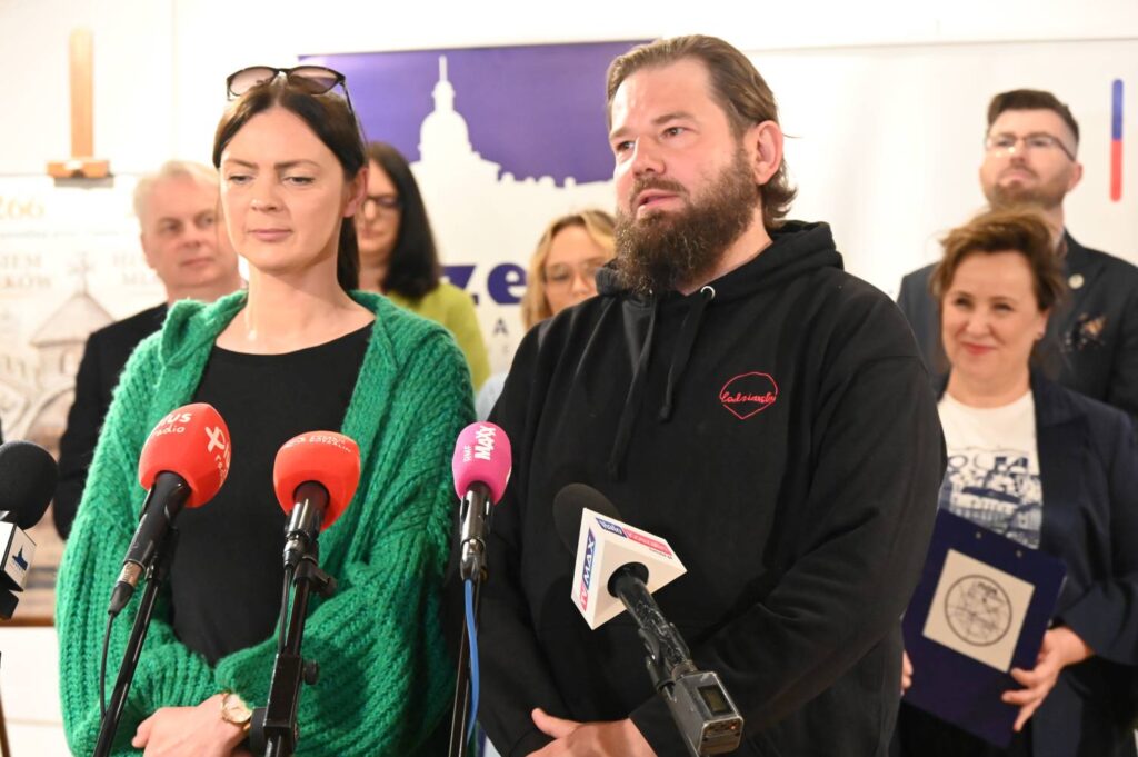 Kobieta w zielonym swetrze i mężczyzna w czarnej bluzie stoją przy mikrofonach podczas konferencji prasowej, w tle widoczni uczestnicy spotkania (kliknięcie powiększy zdjęcie).
