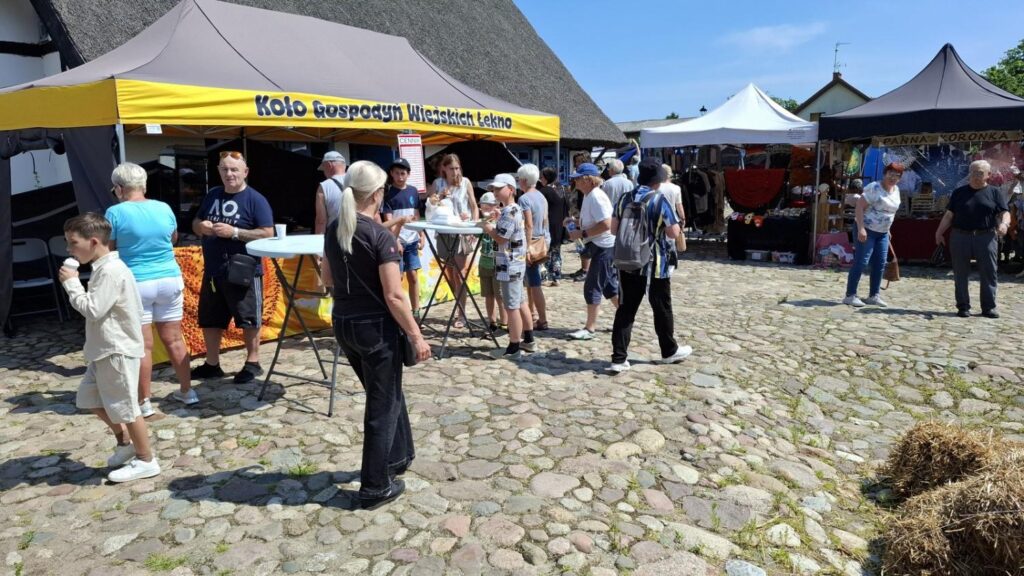 Odwiedzający spacerują między stoiskami jarmarku na terenie skansenu. W tle widoczne są namioty wystawców i zabudowa muzealna. (kliknięcie powiększy zdjęcie).