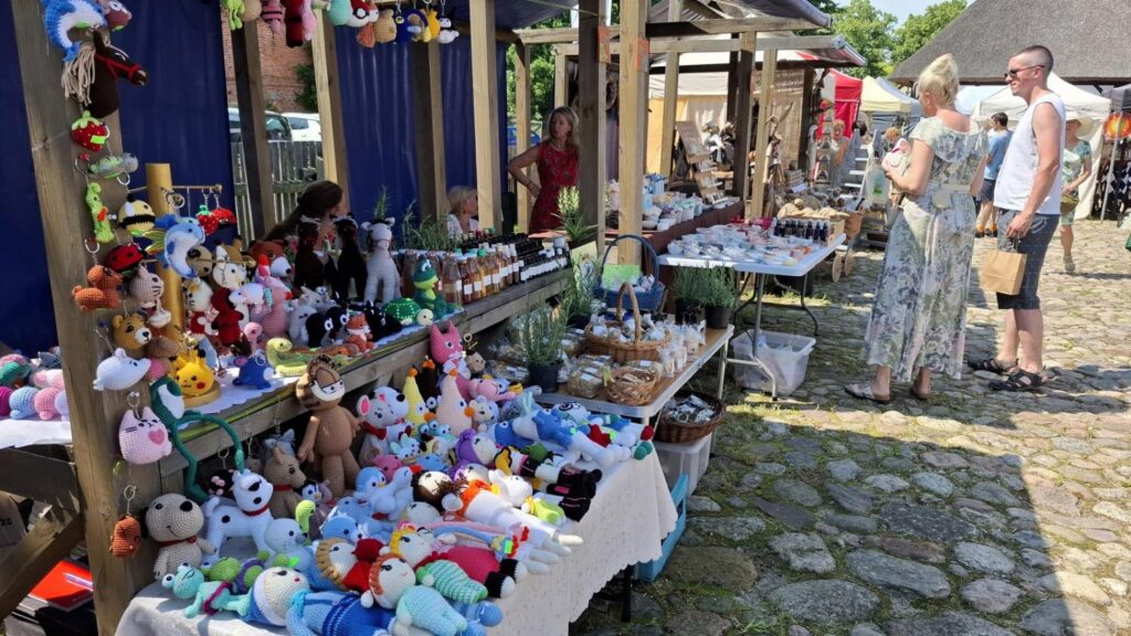 Kolorowe pluszowe zabawki i wyroby rękodzielnicze prezentowane na stoisku podczas jarmarku. Zwiedzający oglądają ofertę wystawcy. (kliknięcie powiększy zdjęcie).