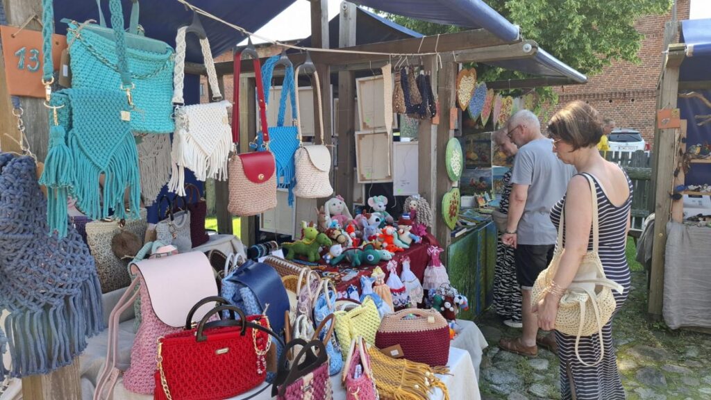 Stoisko z ręcznie wykonanymi torebkami, makramami i kolorowymi zabawkami. Dwie osoby oglądają prezentowane wyroby rękodzielnicze. (kliknięcie powiększy zdjęcie).