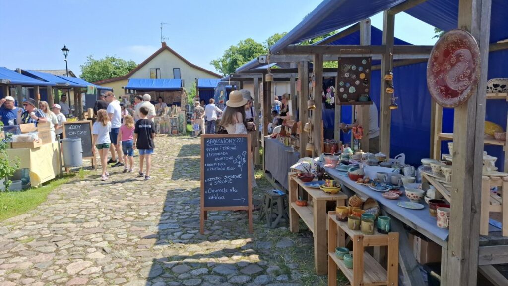 Widok alejki jarmarku z licznymi stoiskami wystawców oraz zwiedzającymi oglądającymi lokalne wyroby artystyczne. (kliknięcie powiększy zdjęcie).