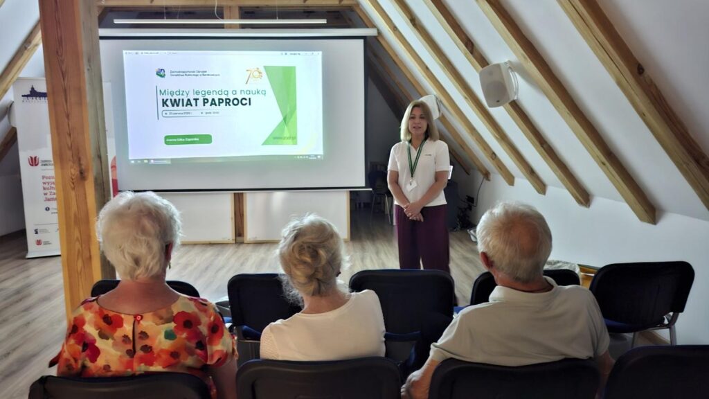 Prelegentka stoi przed ekranem projekcyjnym podczas spotkania w sali na poddaszu muzeum. Na ekranie wyświetlona jest prezentacja poświęcona kwiatowi paproci, a uczestnicy siedzą na widowni. (kliknięcie powiększy zdjęcie).