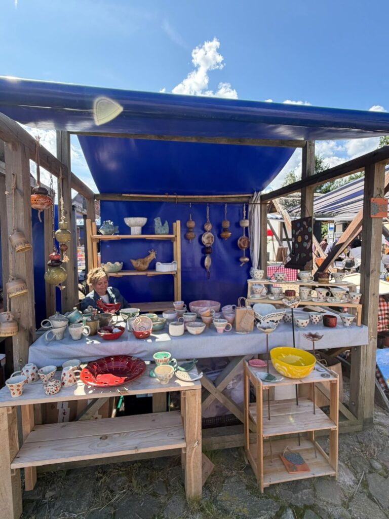 Stoisko z ręcznie wykonaną ceramiką; na drewnianych półkach i stołach prezentowane są kolorowe kubki, miski, talerze, dzbanki i ozdobne naczynia (kliknięcie powiększy zdjęcie..