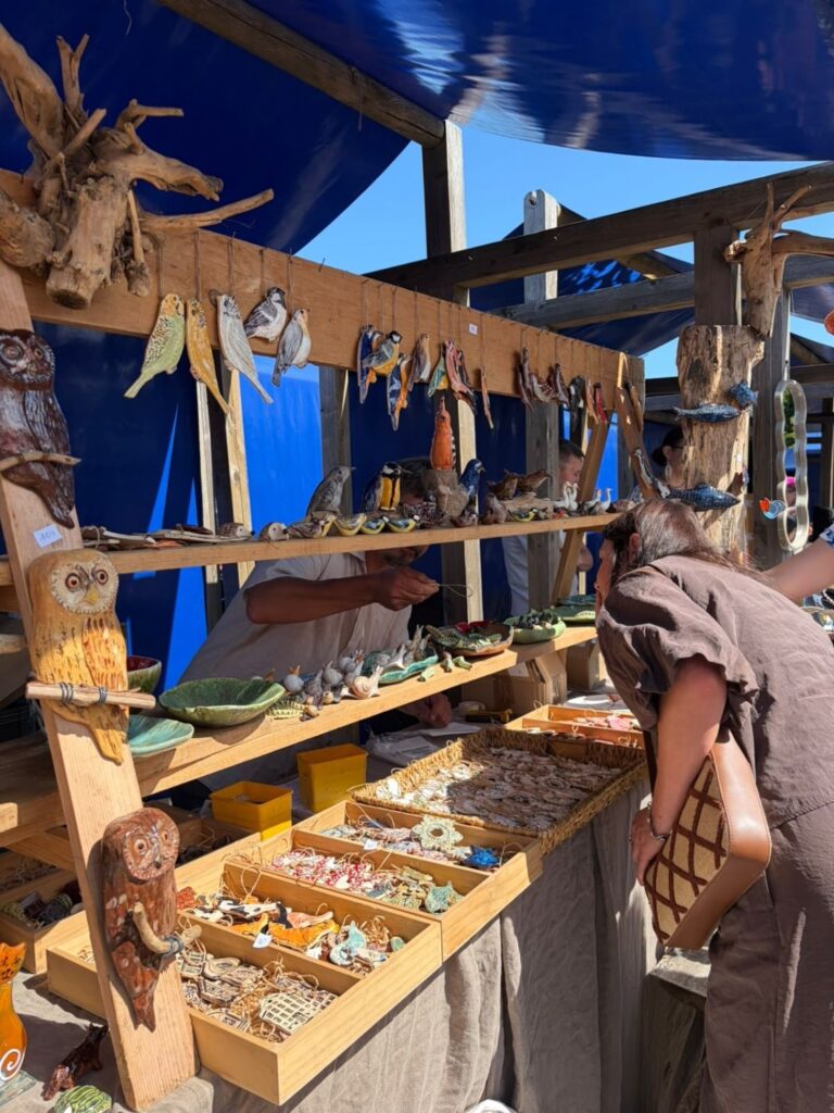 Stoisko z rękodziełem przedstawiającym drewniane i ceramiczne figurki ptaków, sów oraz inne ozdoby; kobieta ogląda oferowane przedmioty. (kliknięcie powiększy zdjęcie).