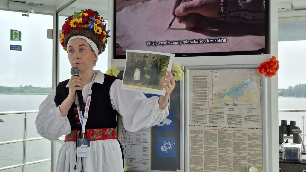 Pracowniczka muzeum w tradycyjnym stroju jamneńskim opowiada o historii Jamna podczas rejsu po jeziorze, trzymając reprodukcję archiwalnej fotografii mieszkańców (kliknięcie powiększy zdjęcie).